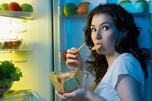 woman eating food in front of open fridge